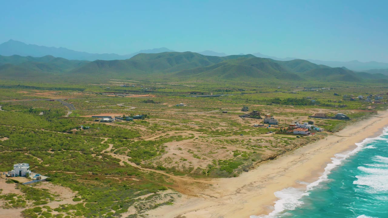 Wide coastal aerial with beach, dunes, homes, and mountains.
