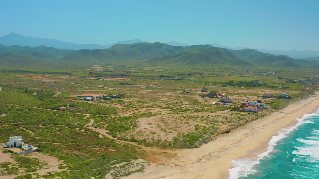 Aerial view across Cerritos with the beach running diagonally beside blue-green water, scrub-covered lots and dirt paths inland, and mountains filling the background, with no discernible people.
