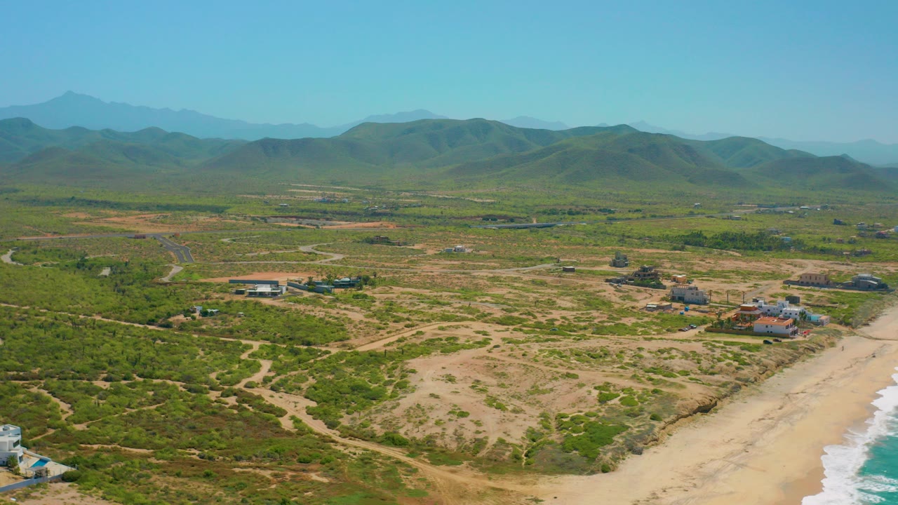 Wide aerial frame over Cerritos, Baja California Sur showing green and tan scrubland, a sandy beach edge with surf at lower right, scattered buildings, distant mountains, and no clearly visible people at this scale.