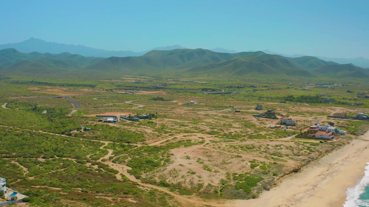 High drone-style view over Cerritos, Baja California Sur showing a broader sweep of green scrub, pale dirt paths, a cluster of buildings near the beach, white surf at the right edge, and distant mountain ridges.