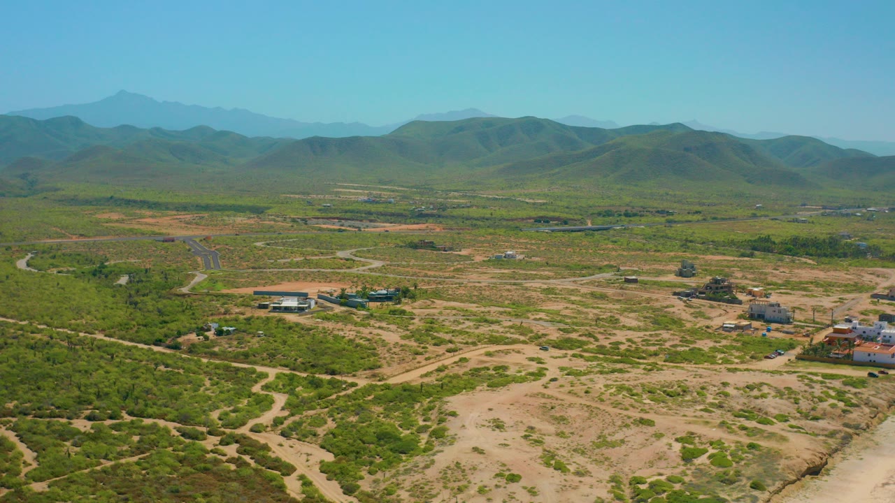Early aerial frame over Cerritos, Baja California Sur, showing tan dirt tracks, green scrub, small buildings, distant hills, and no visible people.