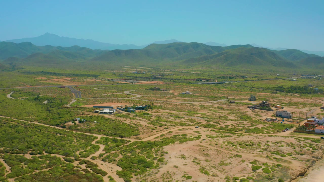 Cerritos, Baja California Sur aerial still with scrub-covered land, pale dirt paths, a few low structures near the right side, hazy hills, and no visible people.