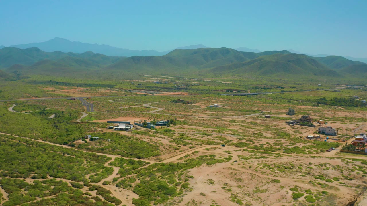 Aerial view across dry open land, green brush, small structures, and distant hills.