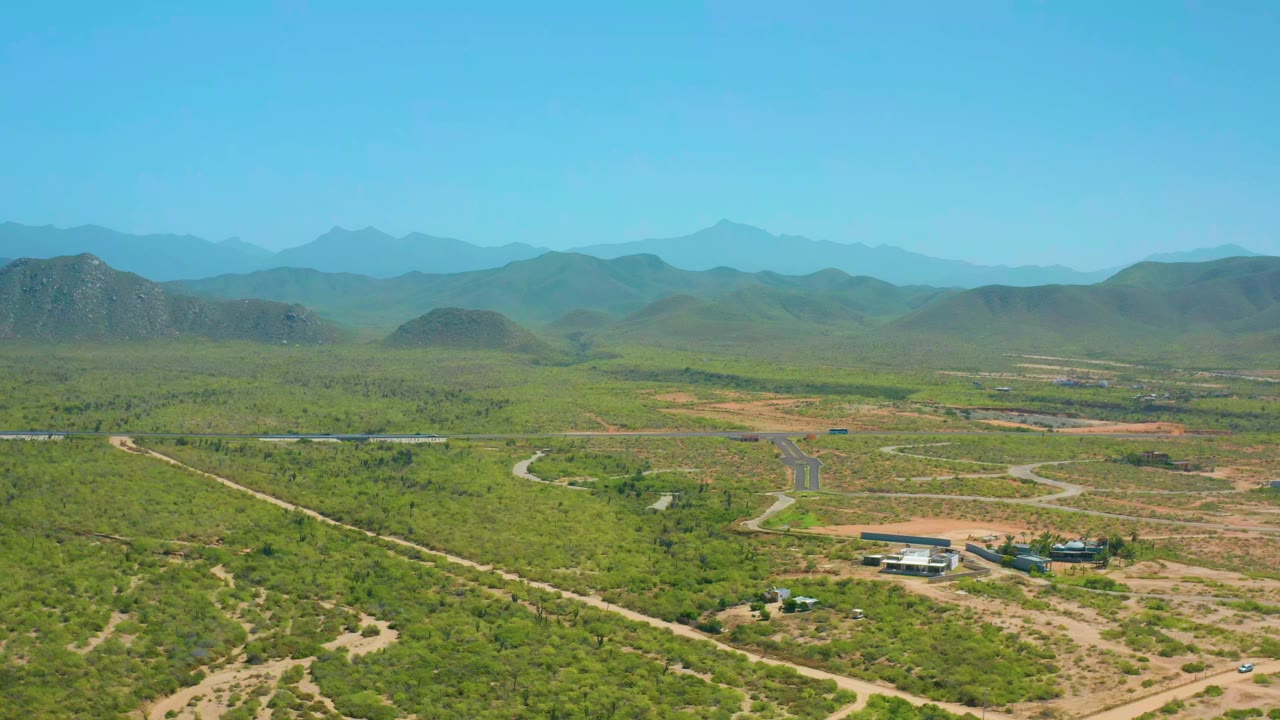 Wide aerial frame in Cerritos, Baja California Sur, with pale roads cutting through green terrain, small structures low in the frame, layered hills across the middle distance, a hazy mountain ridge, and no visible people.