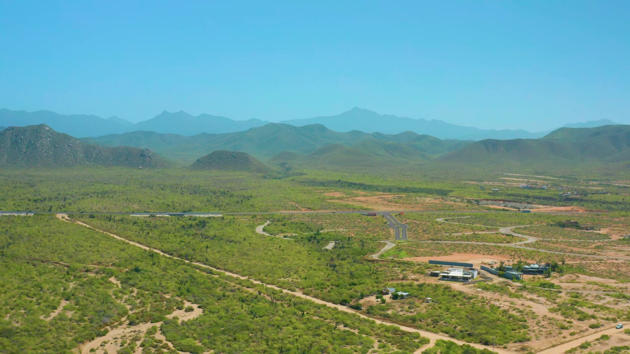 Aerial frame over Cerritos, Baja California Sur, showing the light-roofed compound lower in view, winding dirt tracks through green terrain, layered mountains, and no visible people.