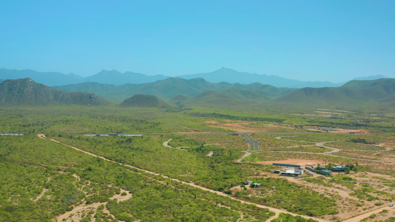 Elevated view across Cerritos, Baja California Sur, with a broad green plateau, pale tracks in the foreground, a paved road in the middle distance, blue mountain layers, and no visible people.