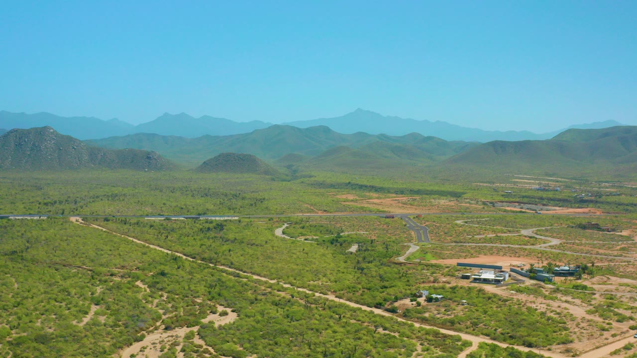 Wide aerial frame over Cerritos, Baja California Sur, showing green scrubland, pale dirt roads, a small building cluster near the lower center, distant hills, and no visible people.