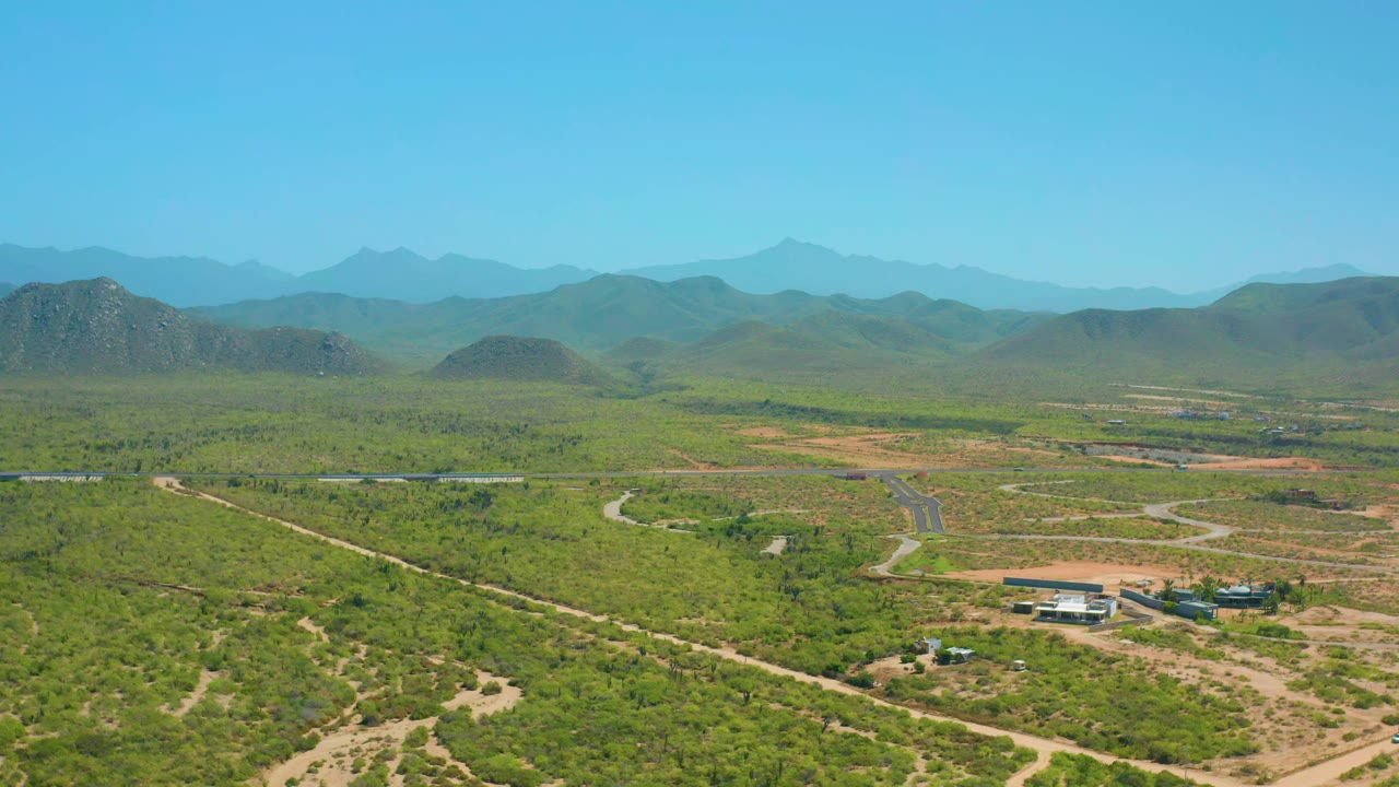 Aerial view in Cerritos, Baja California Sur, with the isolated building cluster lower in frame, winding dirt roads across green terrain, hazy hills beyond, and no visible people.
