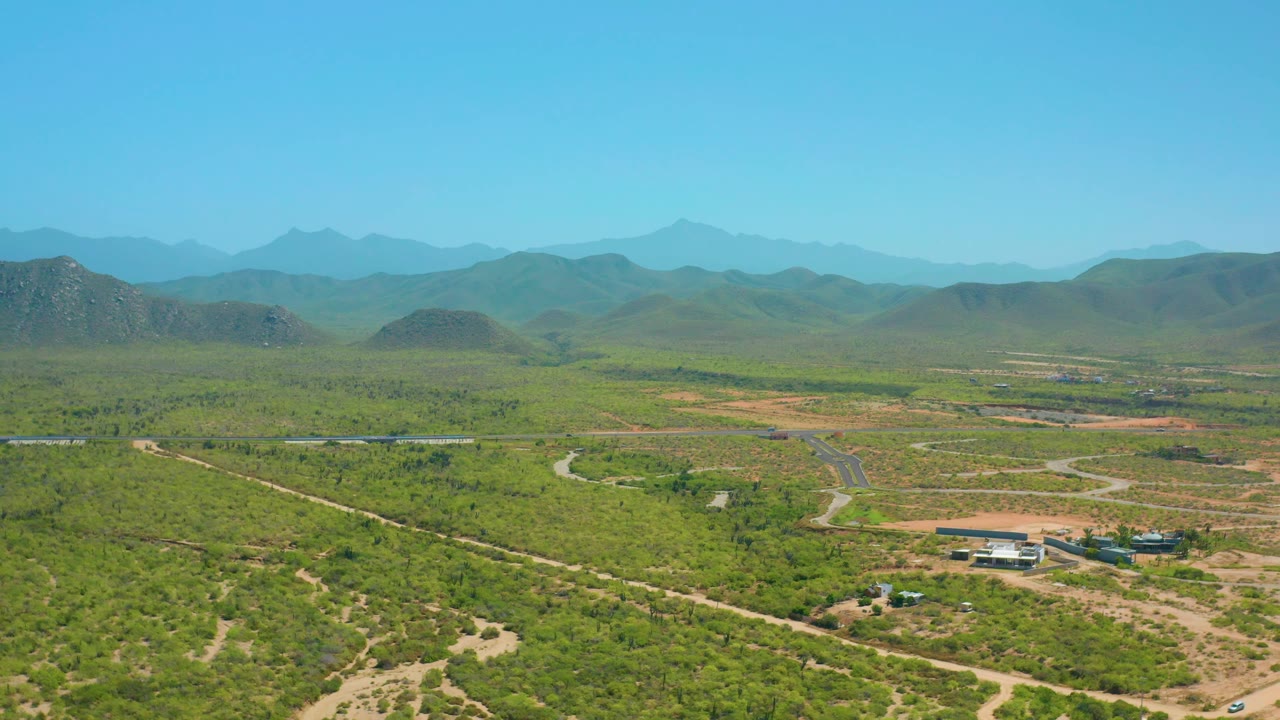 The Cerritos, Baja California Sur view shows sparse green terrain broken by light dirt paths, a small cluster of buildings, layered hills, and no visible people.