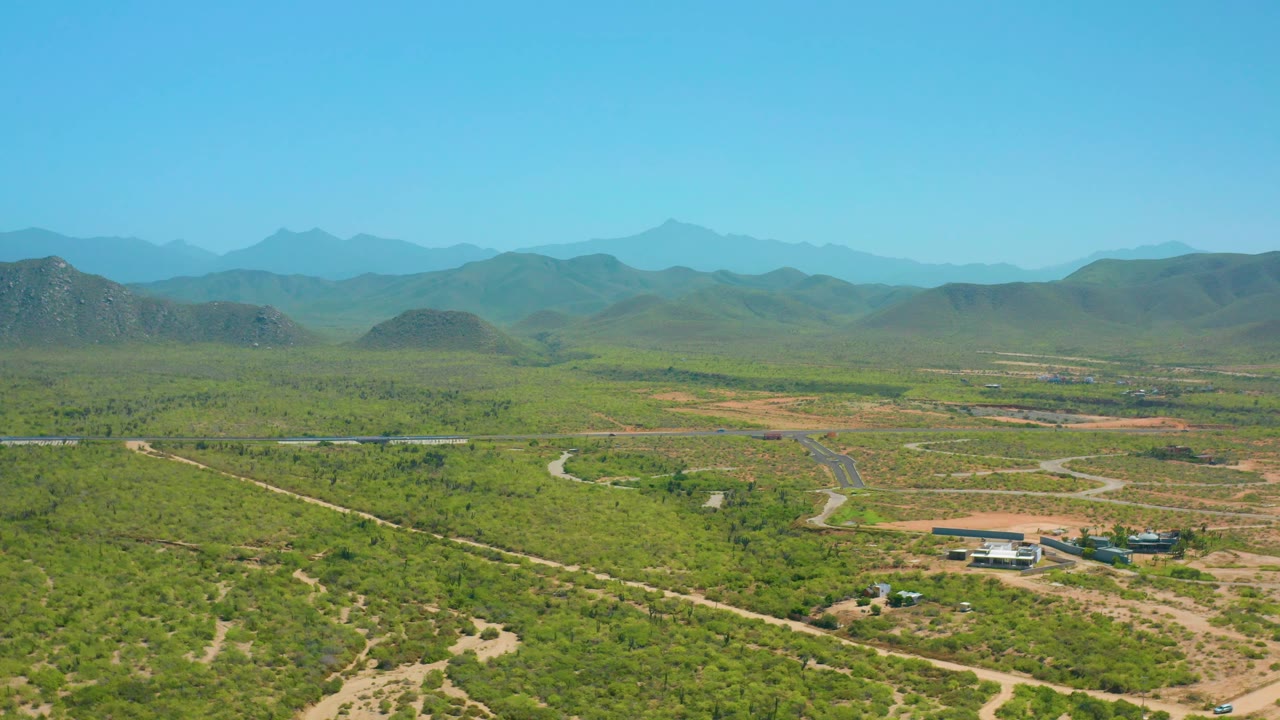 Mid-clip aerial view near Cerritos with a paved road cutting through scrubland, winding tracks in the foreground, distant mountain ridges, and no visible people.