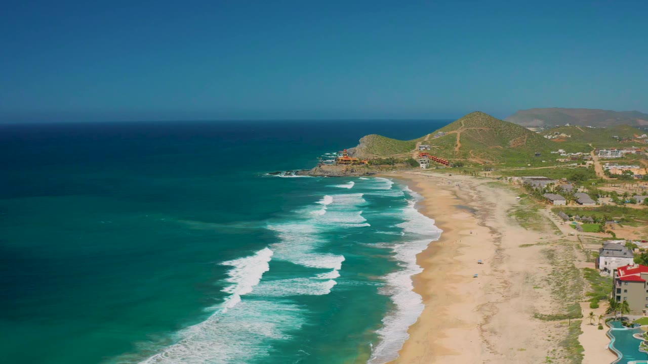 Wide aerial frame of Cerritos coastline showing foam bands farther down the beach, open sand through the center, hillside terrain beyond, and no visible people.