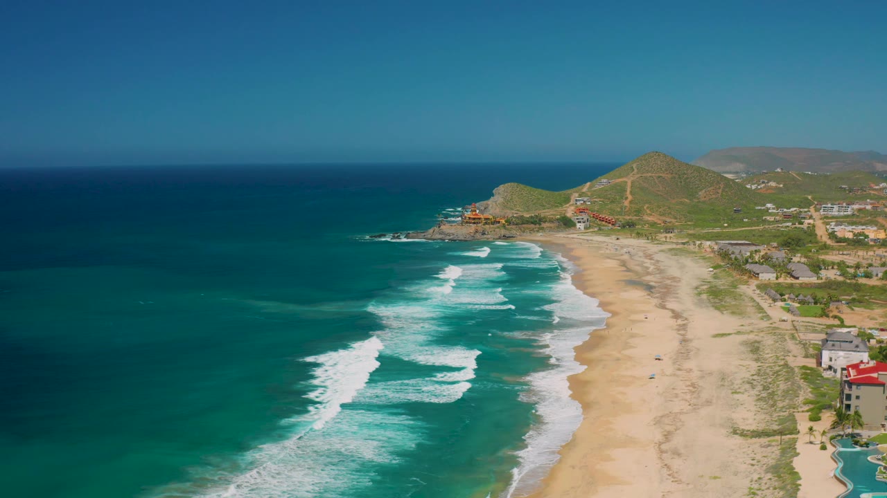 Drone-height view over Cerritos with darker blue water offshore, white wave sets near shore, a tan beach, hillside roads, coastal buildings, and no visible people.