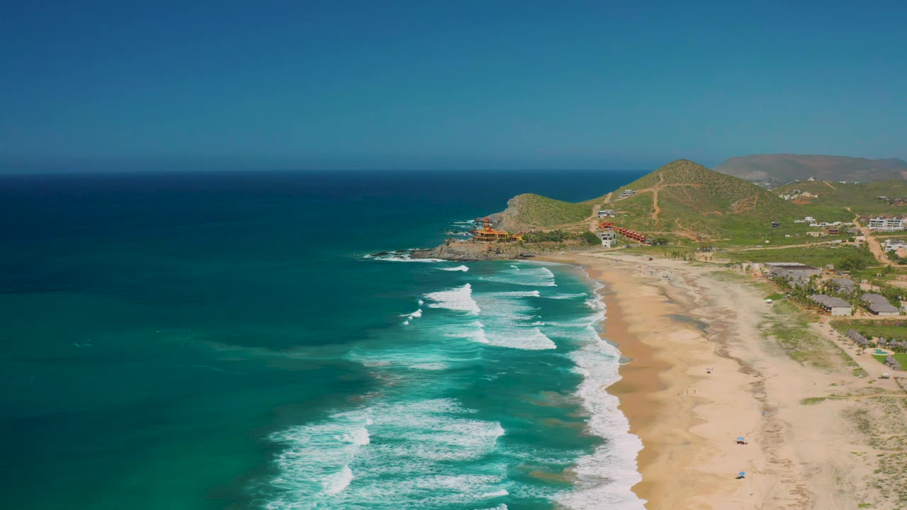 Second aerial frame of Cerritos coastline with deeper blue water near the horizon, turquoise shallows, diagonal surf lines, tan beach, inland hills, and no visible people.