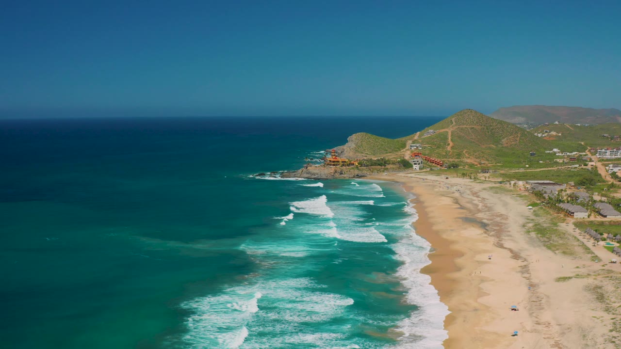 Cerritos aerial view with white wave sets crossing the middle of the frame, open sand on the right, scattered beachside structures, green hills behind, and no visible people.