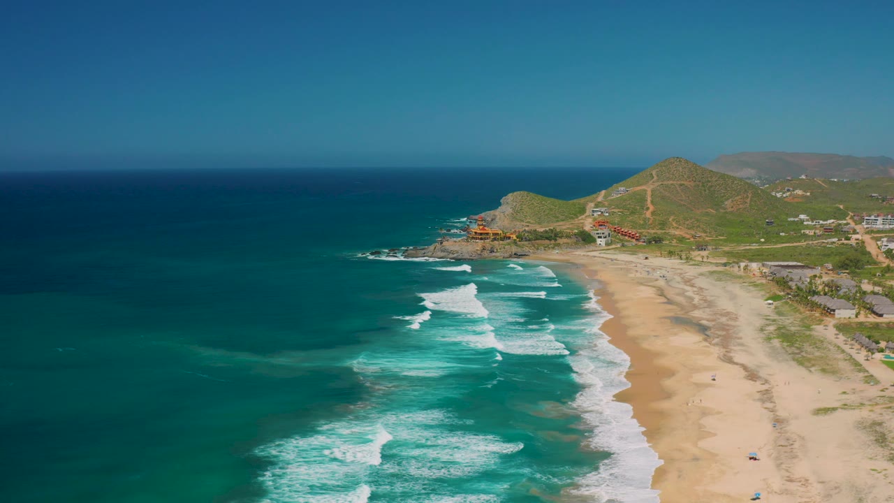 Mid-clip aerial frame over Cerritos showing a broad band of ocean, foamy surf curling toward the beach, light sand, a few small buildings near the hill, and no visible people.