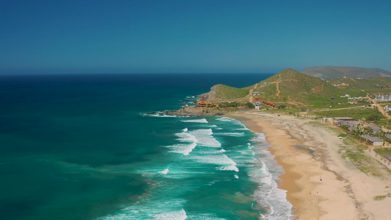 Aerial frame over Cerritos, Baja California Sur with turquoise water on the left, white surf lines reaching a broad sand beach, green hills in the distance, and no visible people.
