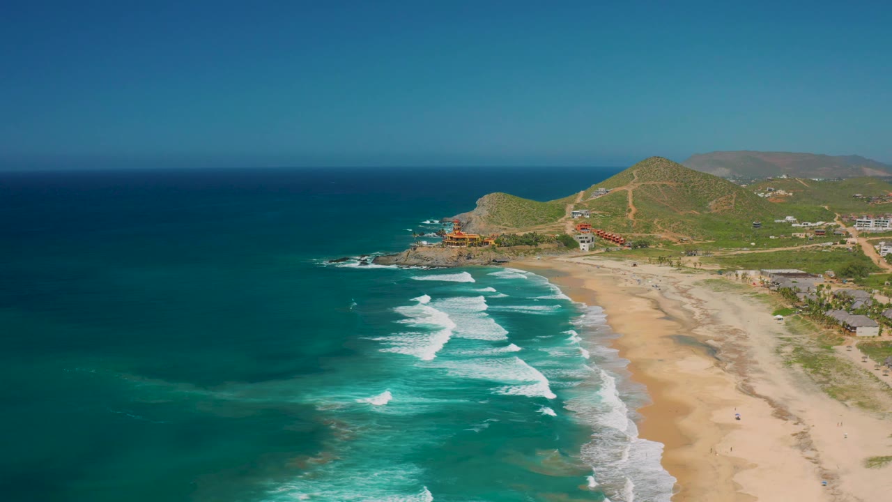 Cerritos, Baja California Sur shoreline seen from above, with whitecaps advancing in staggered rows, a pale sand beach on the right, hillside roads and buildings beyond, and no visible people.