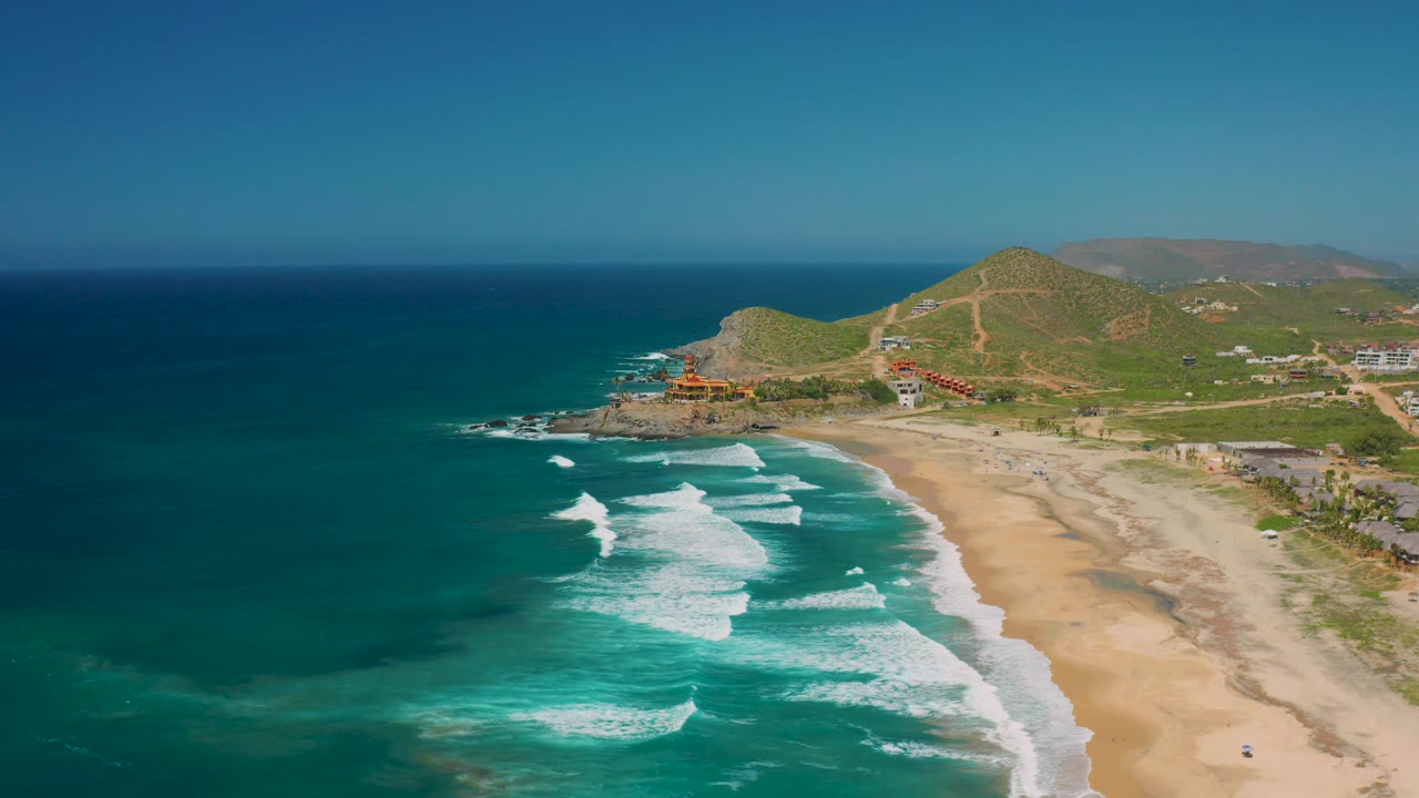 Aerial view of Cerritos, Baja California Sur with deep blue sea, white surf bands, long sandy beach, green hills, scattered buildings, and no visible people.