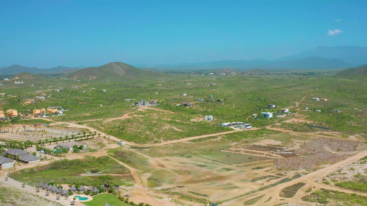 Opening aerial frame over Cerritos, Baja California Sur, with green scrubland, tan dirt roads, scattered low buildings, distant hills, hazy mountains, bright blue sky, and no visible people.