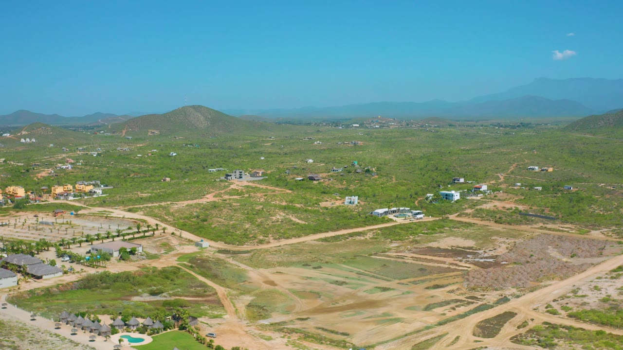 Third frame over Cerritos, Baja California Sur, with dirt roads crossing green and tan ground, scattered houses or small structures, rounded hills, a hazy mountain line, small clouds, and no visible people.