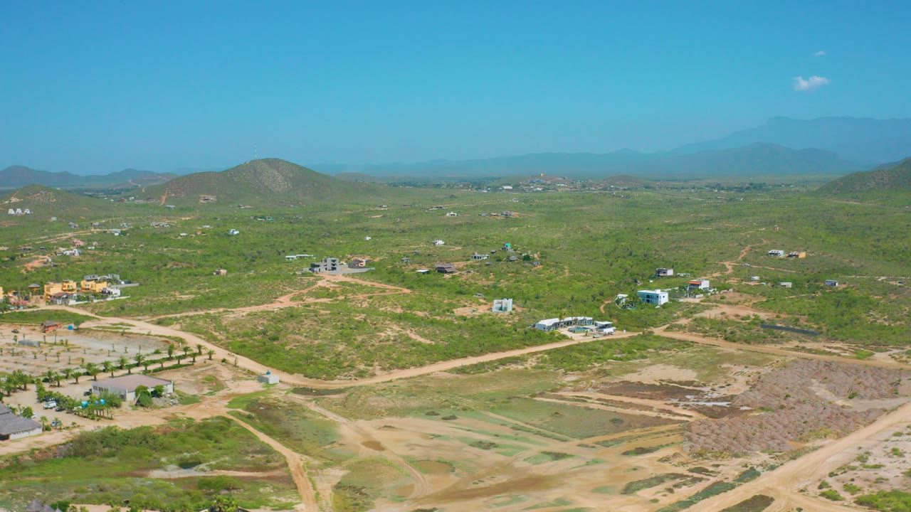 Opening aerial frame over Cerritos, Baja California Sur, Mexico, showing green scrubland crossed by pale dirt roads, scattered buildings, low hills, distant mountains, and no visible people.