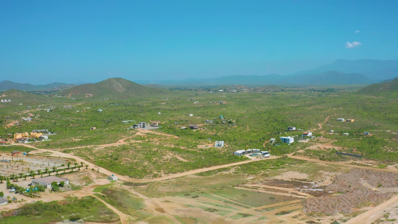 Aerial view across scrubland with a central dirt road, buildings, hills, and distant ridge.