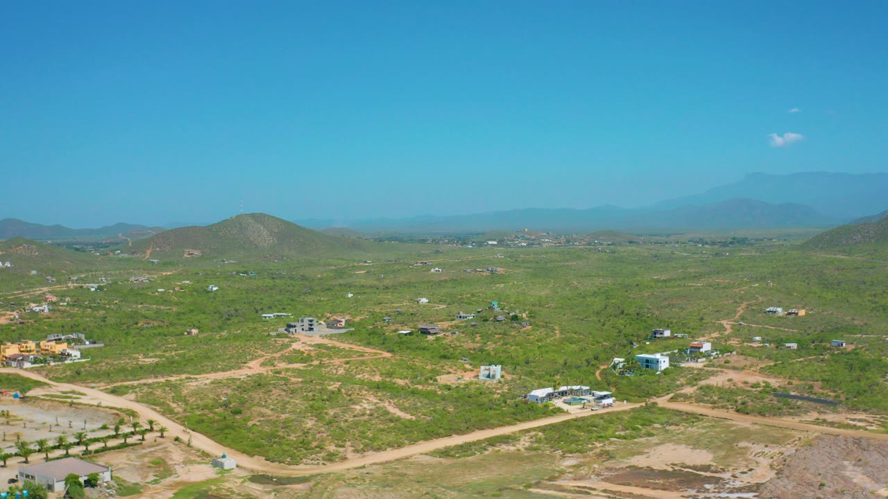 Opening aerial frame over Cerritos, Baja California Sur with green scrubland, pale dirt roads, scattered buildings, low hills, distant mountains, blue sky, and no visible people.