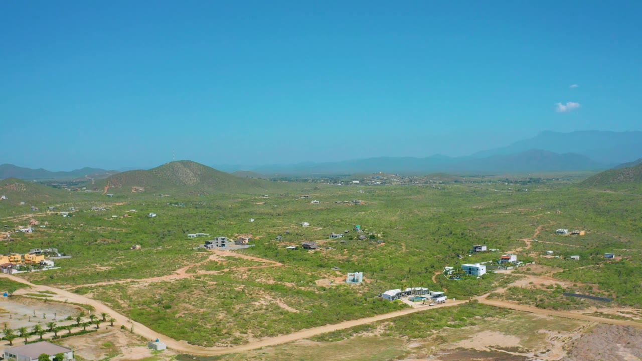 Cerritos aerial frame with a broad blue sky above green scrubland, winding dirt roads, small light buildings, low hills, distant mountain silhouettes, and no visible people.