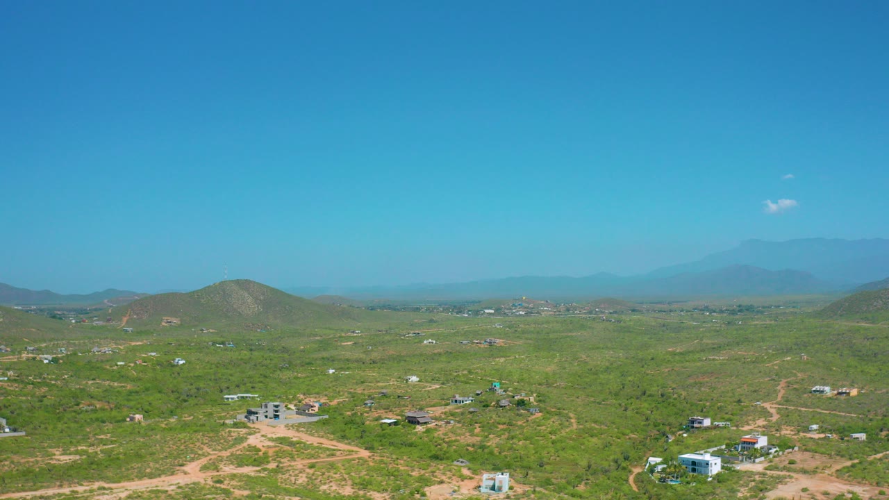Opening frame of Cerritos, Baja California Sur showing green scrubland, tan dirt tracks, scattered small buildings, rounded hills, hazy mountains, and no visible people.