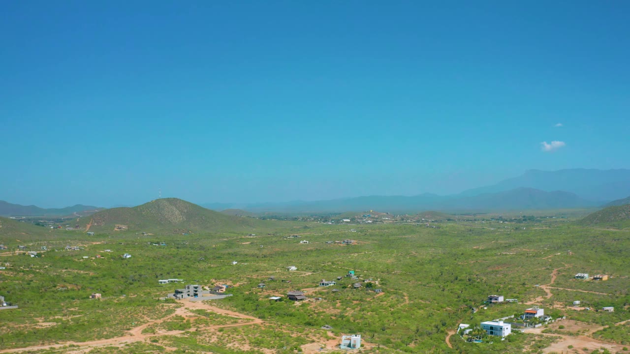 Aerial landscape with scattered buildings, dirt tracks, green brush, and distant mountains.