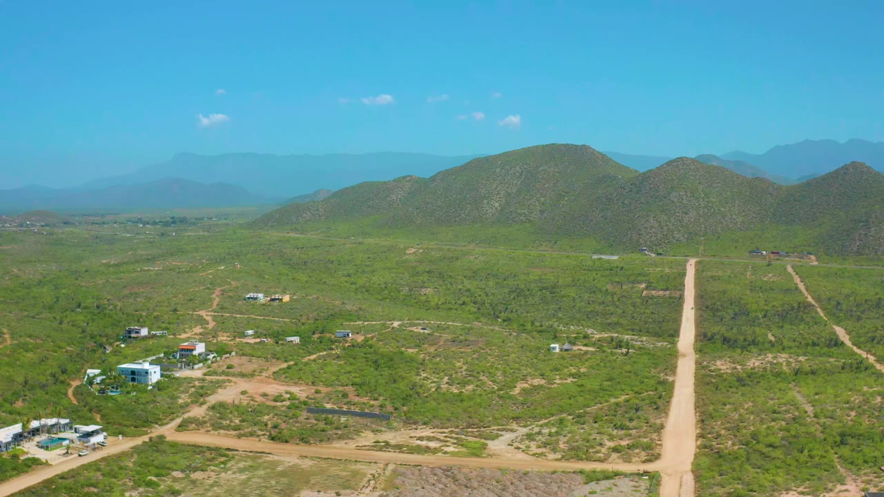 Aerial view in Cerritos, Baja California Sur showing a straight pale dirt road crossing green scrubland toward rounded hills, with scattered buildings and no visible people.