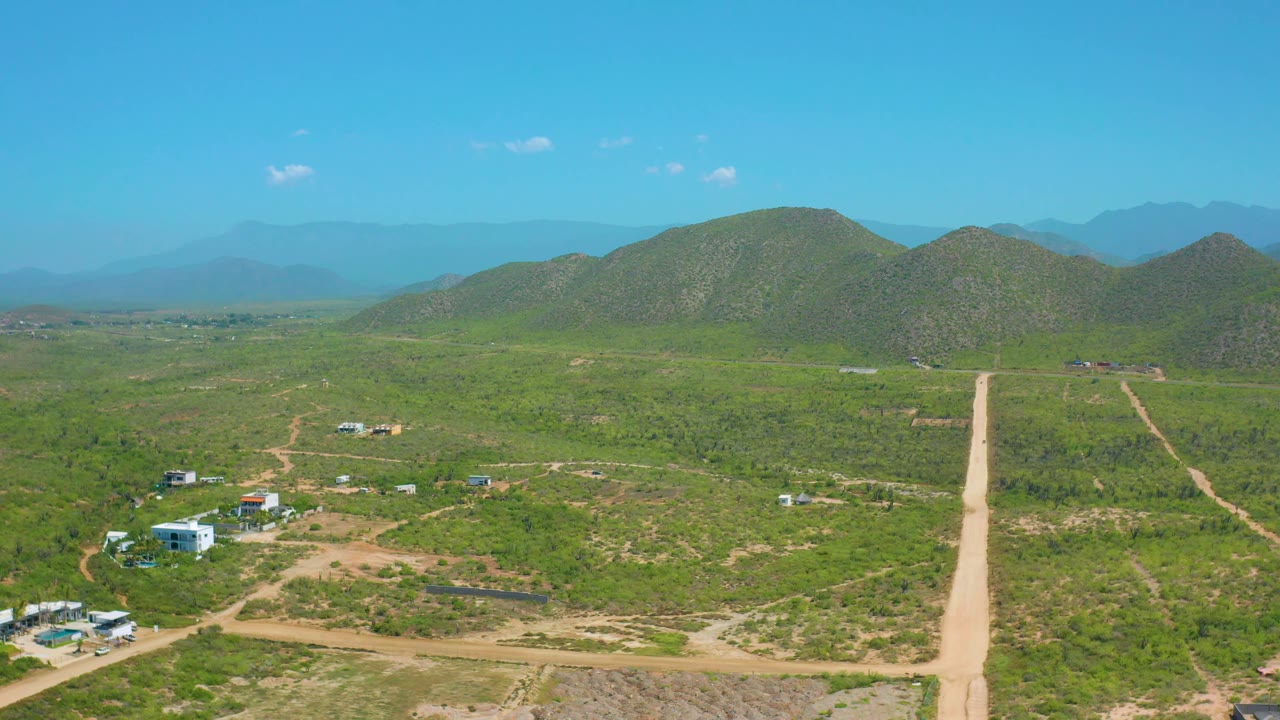 Aerial view in Cerritos, Baja California Sur with a straight dirt road centered in the frame, green brush on both sides, scattered buildings, distant mountains, and no visible people.