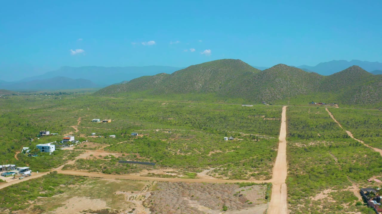 Wide aerial view of a straight dirt road leading across green land toward rounded hills.