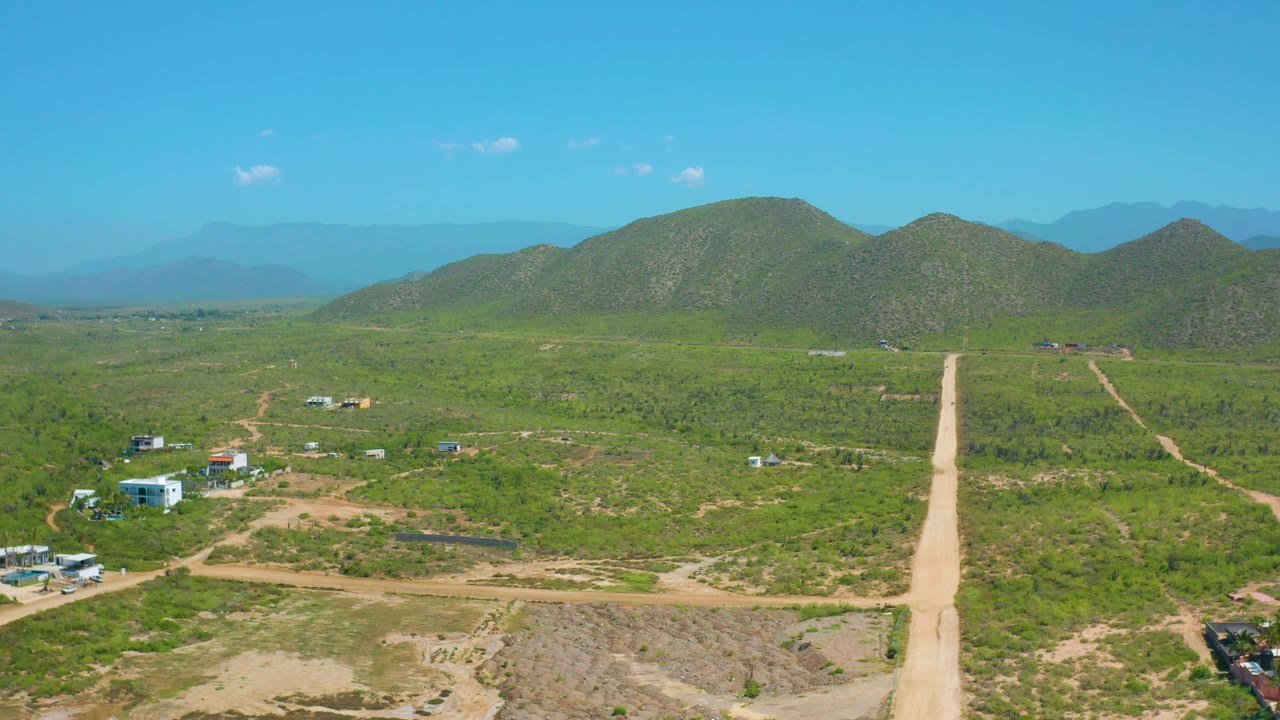 At 0:02, the Cerritos aerial view shows pale intersecting dirt roads, sparse homes, green terrain, and low hills under a clear blue sky with no visible people.