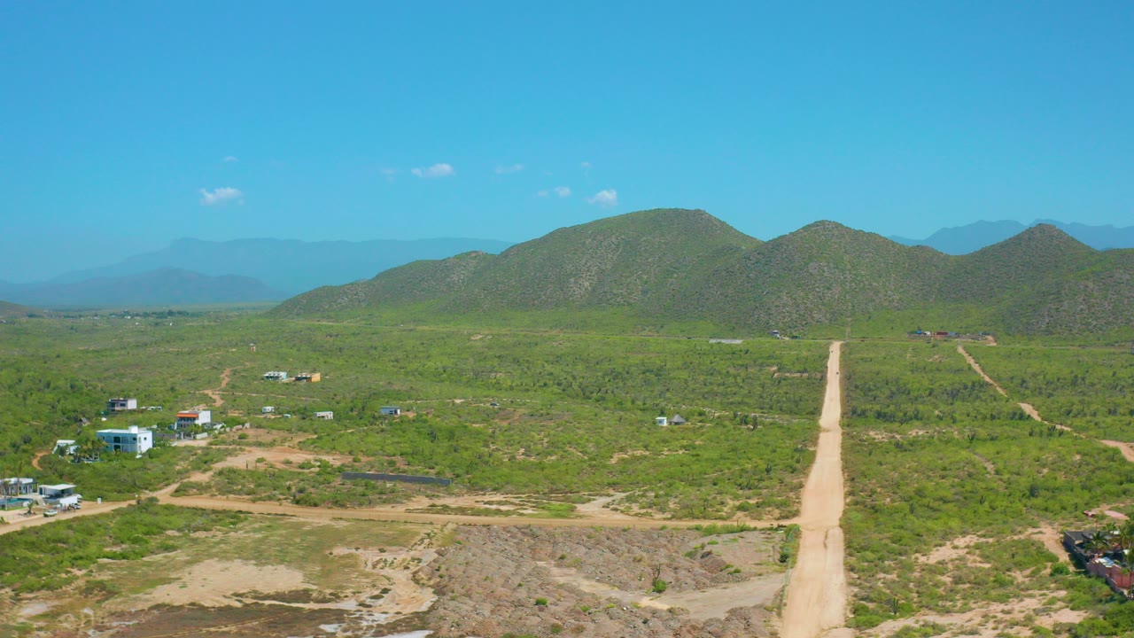 Opening frame of a pale dirt road in Cerritos, Baja California Sur, running through green rural land toward rounded hills, with scattered buildings and no visible people.