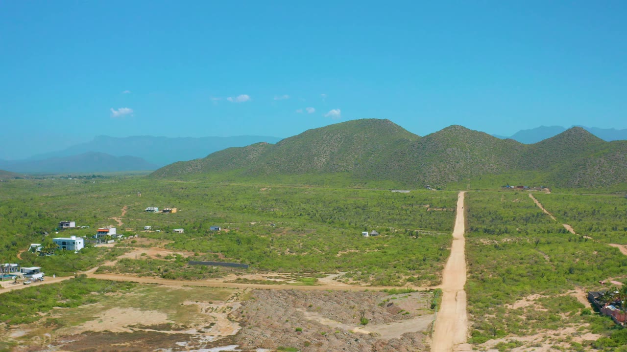 At 0:02, the aerial view shows the pale road crossing from foreground toward the hills in Cerritos, with sparse structures on both sides and no visible people.