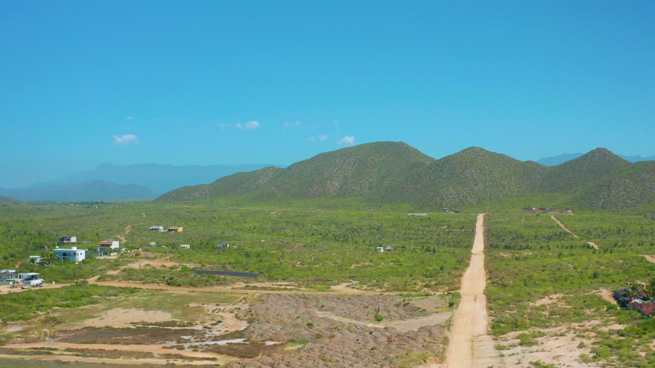 Aerial frame in Cerritos, Baja California Sur showing a pale dirt road running toward green hills, scattered small buildings, scrubby green land, blue sky, and no visible people.