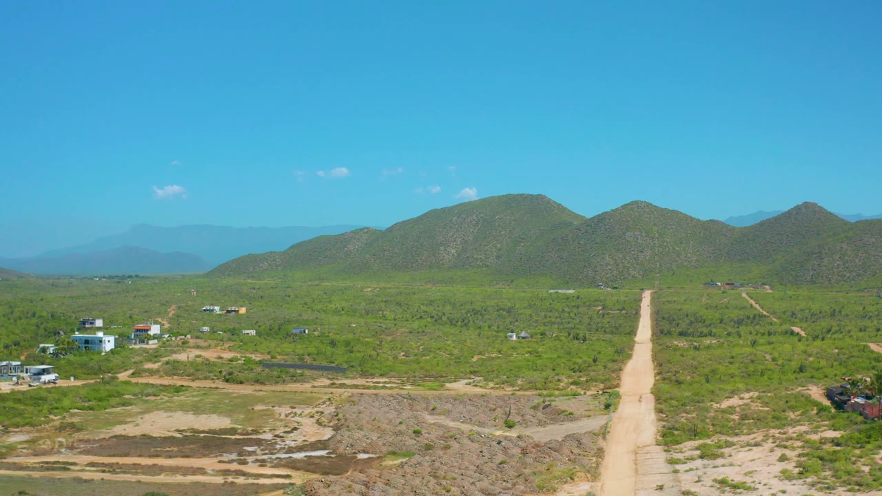 Cerritos, Baja California Sur aerial frame showing the road crossing open green terrain, low buildings near the left and right edges, rounded hills across the horizon, and no visible people.