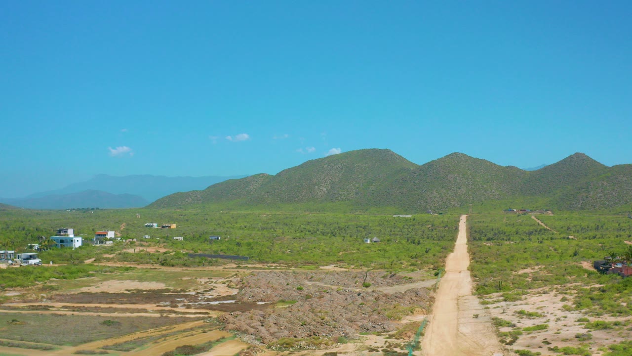 Wide aerial frame in Cerritos, Baja California Sur with a pale dirt road centered toward green hills, scattered low buildings, scrub vegetation, and no visible people.