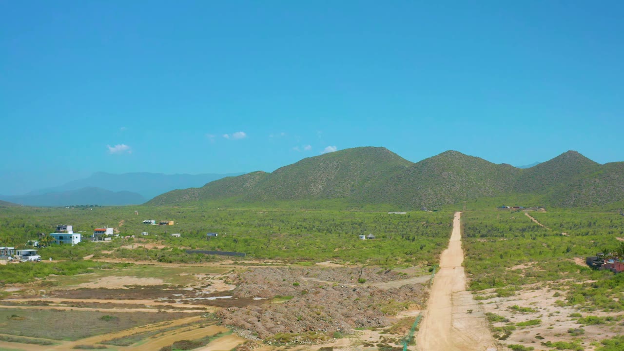 Aerial road view with green plants, small buildings, and low mountains in the distance.