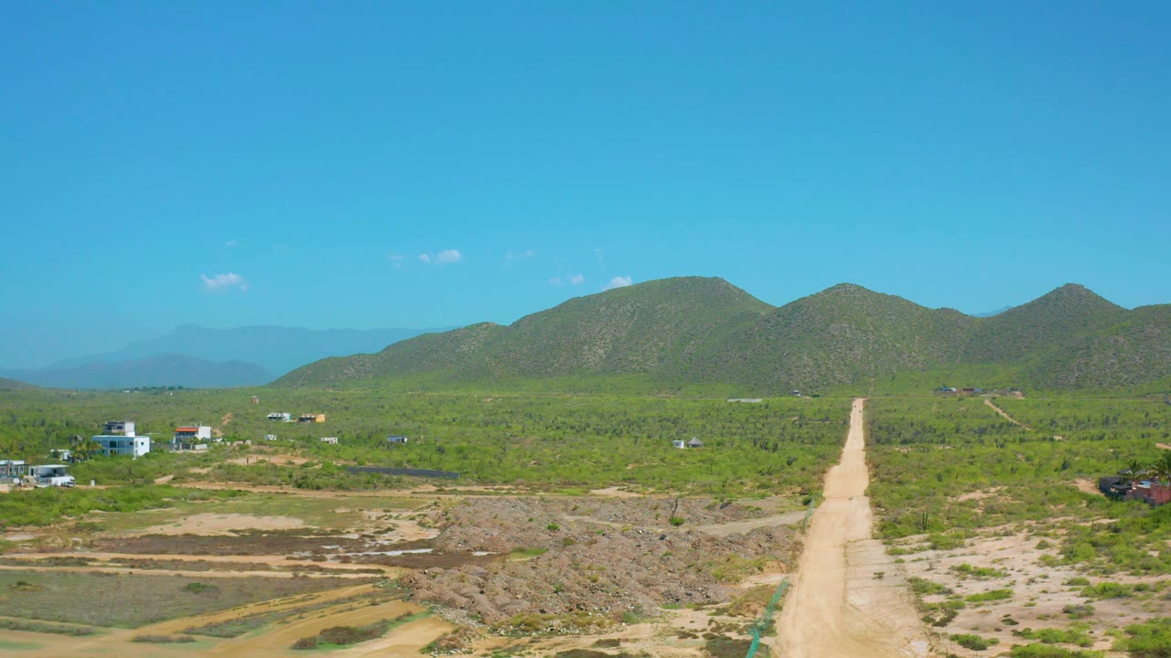 Cerritos, Baja California Sur aerial frame with the dirt road running from foreground earth toward rounded green mountains, with scattered buildings and no visible people.