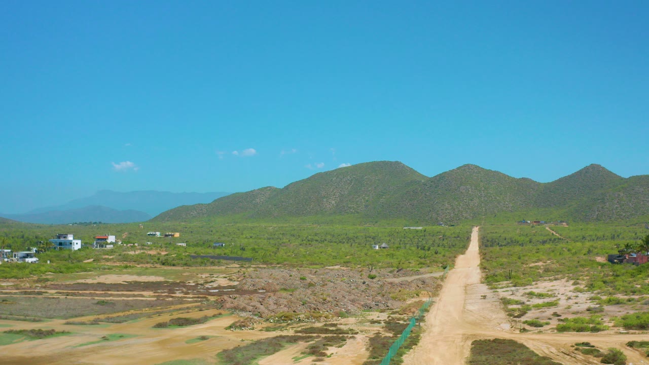 Opening frame of Cerritos, Baja California Sur with a pale dirt road cutting through dry graded ground toward green mountains, small buildings at the edges, and no visible people.