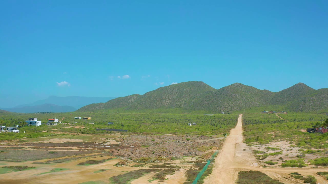 Cerritos aerial frame with the dirt road running up the center, dry lots and scrub in the foreground, distant small buildings, green hills, and no visible people.
