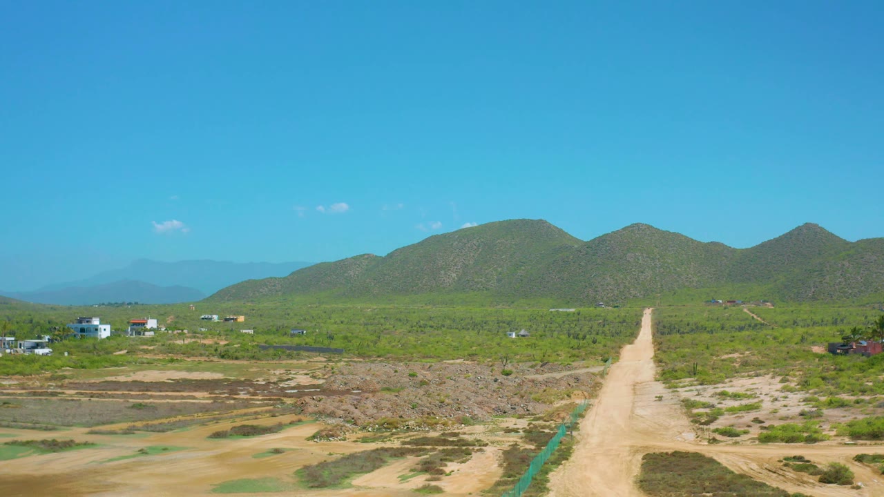 The frame shows Cerritos rural terrain with a pale road, low scrub, scattered small structures on both sides, rounded green mountain slopes, and no visible people.