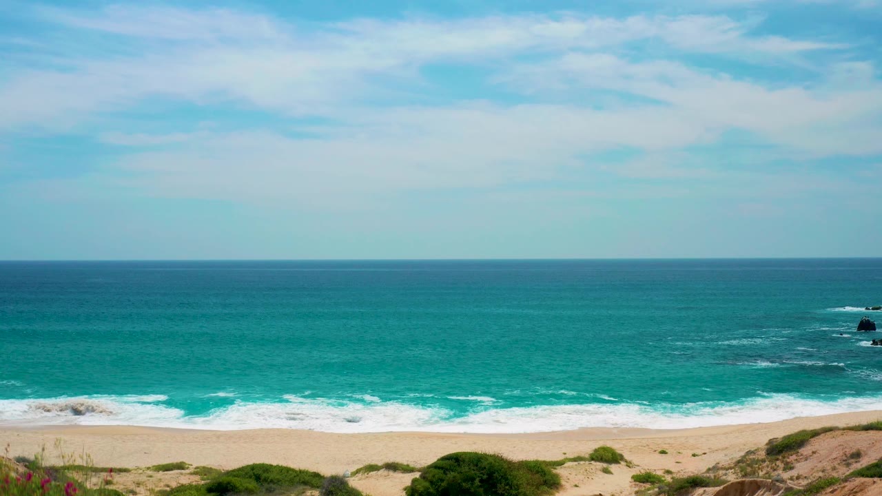 At 0:03, the frame centers on the open ocean and horizontal surf bands, with sand and low vegetation at the bottom edge of Secrets Marquis Los Cabos, Mexico, and no visible people.