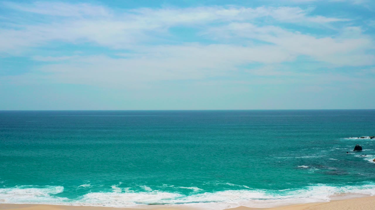 At Secrets Marquis Los Cabos, Mexico, turquoise water rolls toward a thin sandy beach with offshore rocks near the right side and no visible people.