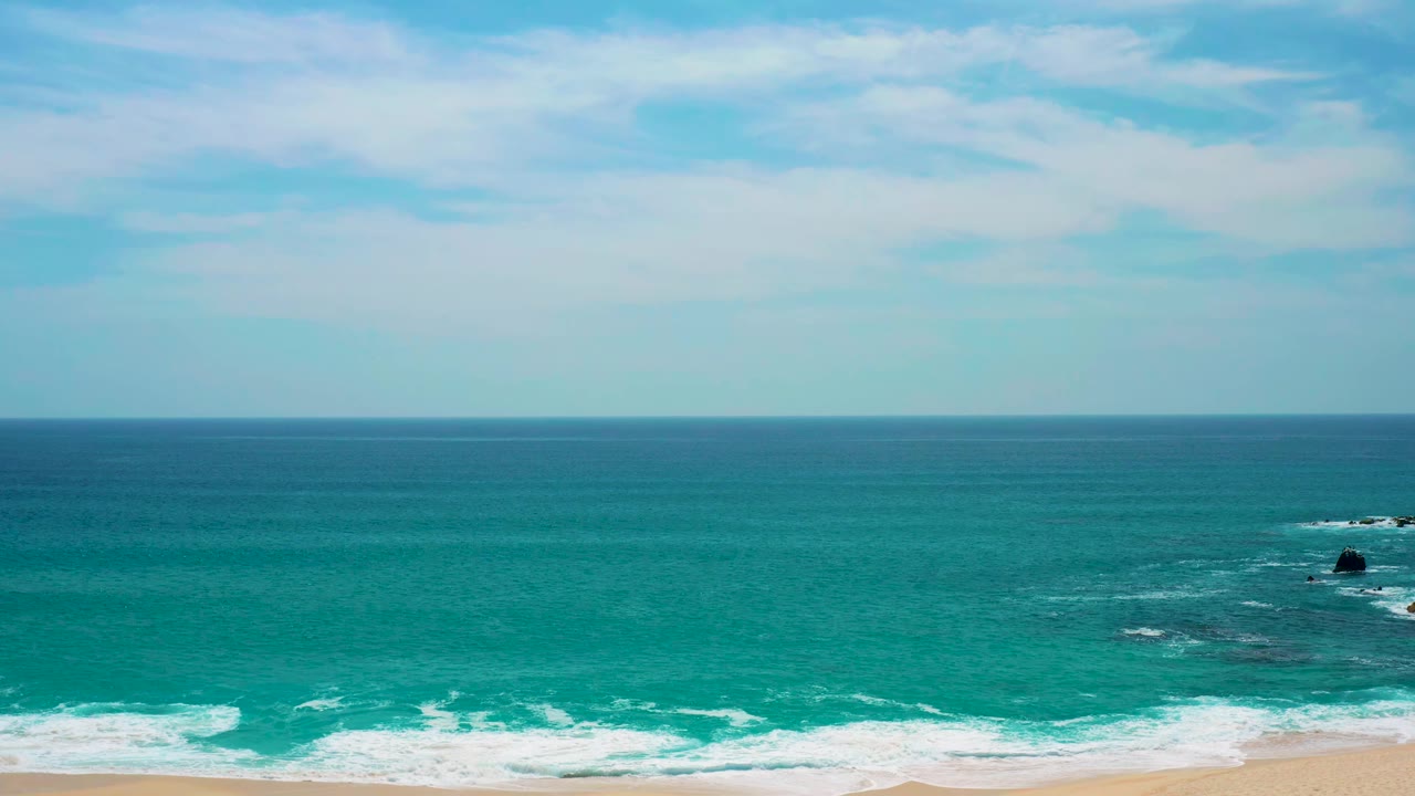 At Secrets Marquis Los Cabos, Mexico, the surf spreads along the lower edge while a dark offshore rock sits in blue-green water beneath a level horizon and no visible people.