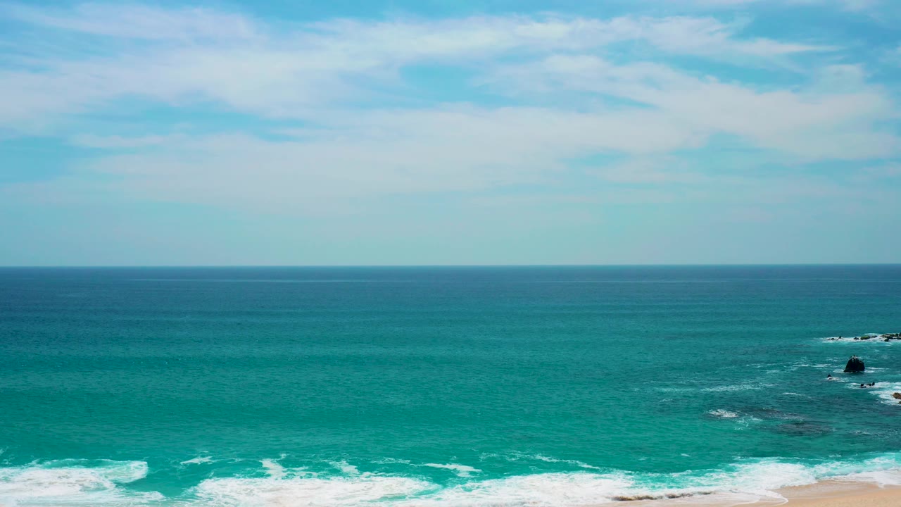 At Secrets Marquis Los Cabos, Mexico, the shoreline surf runs across the bottom while small rocks appear toward the far right in open turquoise water with no visible people.