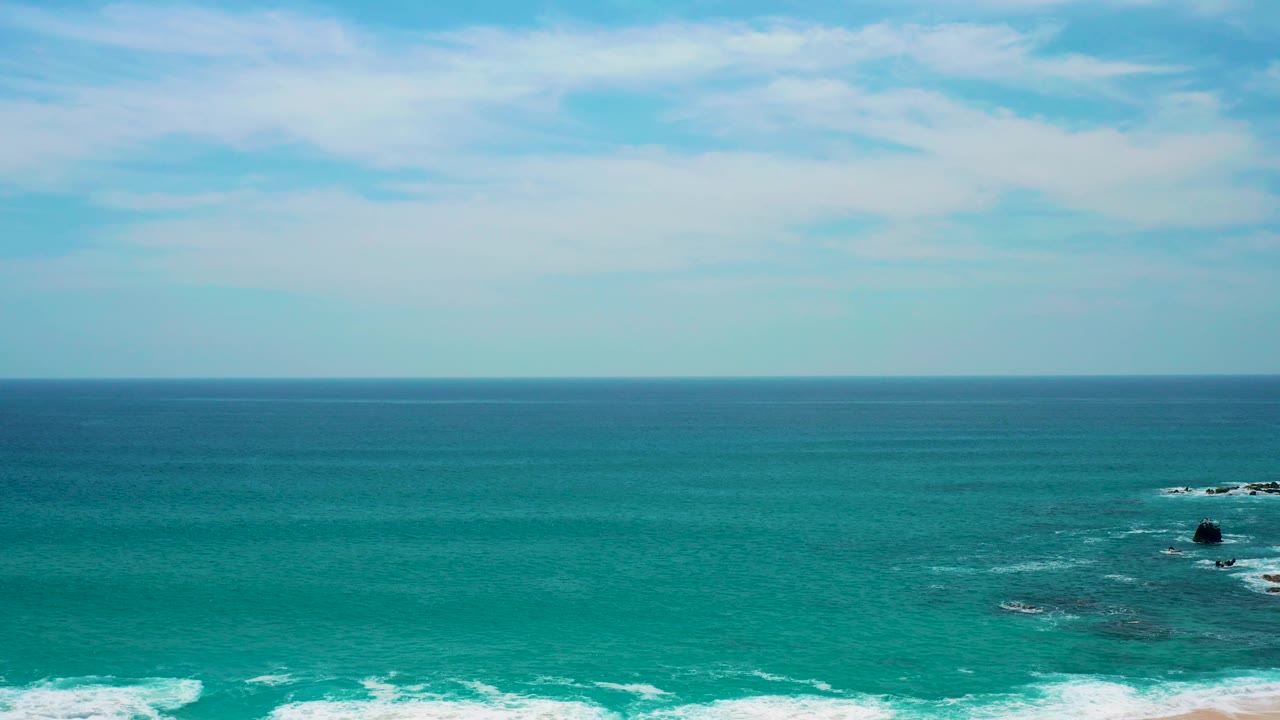 At Secrets Marquis Los Cabos, Mexico, a dark rock sits low in the frame beyond the surf, with layered turquoise water, pale clouds, and no visible people.