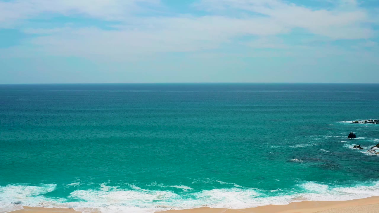 One second into the clip, the Secrets Marquis Los Cabos shoreline shows white wave lines closer to the sand, dark patches below the water, offshore rocks to the right, and no visible people.